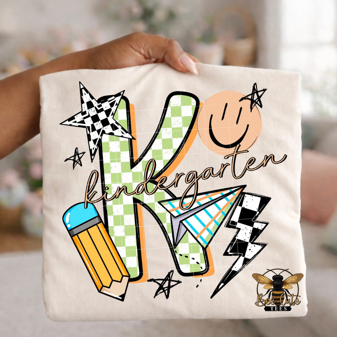 Folded white-colored unisex T-shirt held up by a hand, featuring a colorful "kindergarten" graphic. The design includes green checkered number four, bold black lettering, peach script, a pencil, paper airplane, smiley face, lightning bolt, stars, and playful doodles. A fun, retro-inspired back-to-school design perfect for fourth grade teachers and students.
