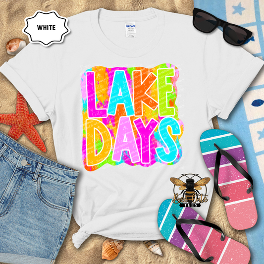 White t-shirt with 'Lake Days' design, sunglasses, and flip-flops on a beach-themed background.