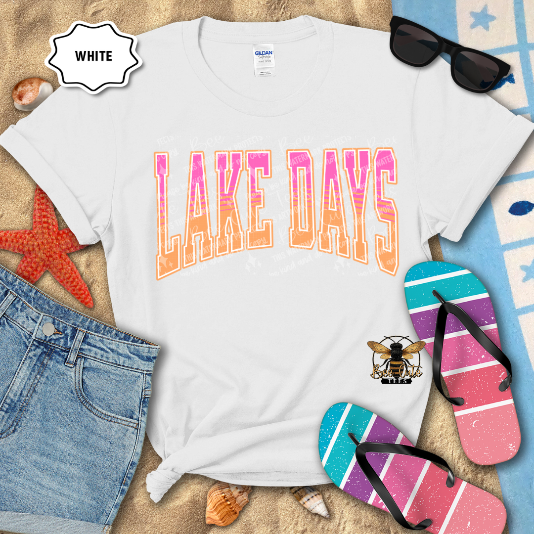 White t-shirt with 'LAKE DAYS' text, sunglasses, flip-flops, and denim shorts on a sandy background.
