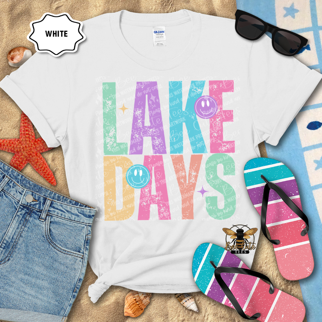 White t-shirt with 'LAKE DAYS' text on a beach-themed background with flip-flops and sunglasses.