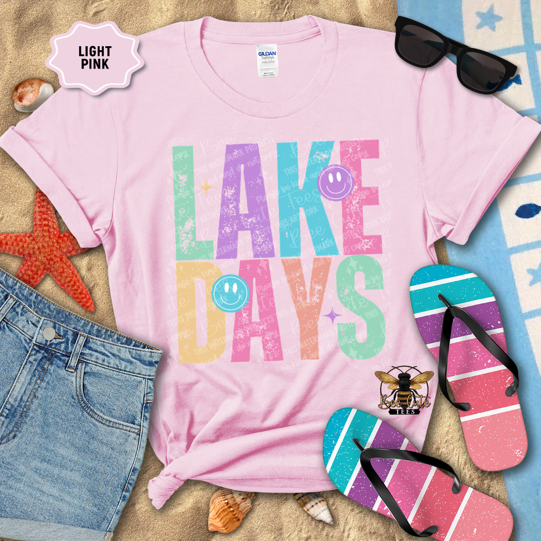 Pink t-shirt with 'Lake Days' text surrounded by beach accessories on a sandy background
