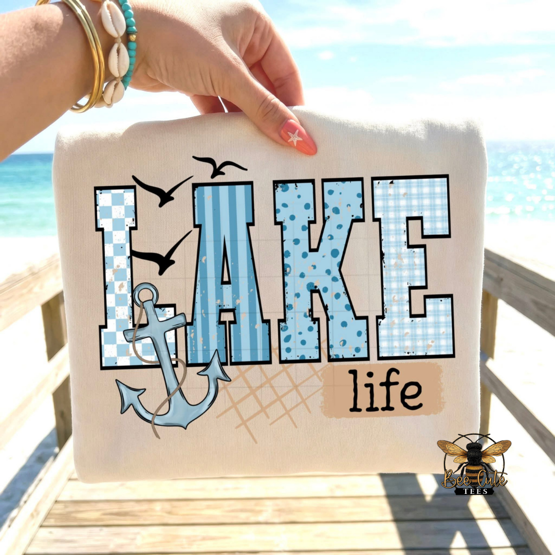 Beige t-shirt with 'LAKE life' design held by a hand on a wooden dock with ocean view.