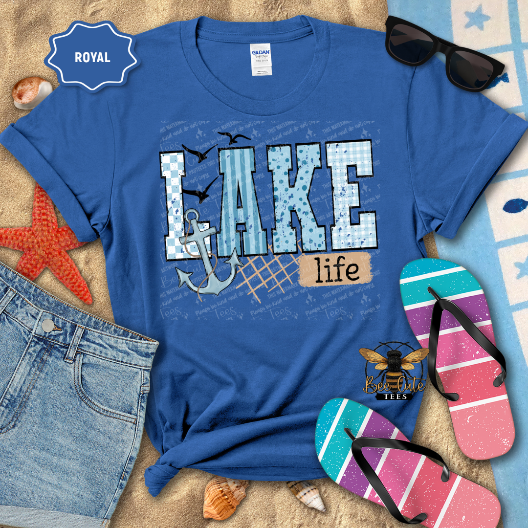 Blue t-shirt with 'Lake life' design on a beach-themed background with sunglasses, flip-flops, and denim shorts.