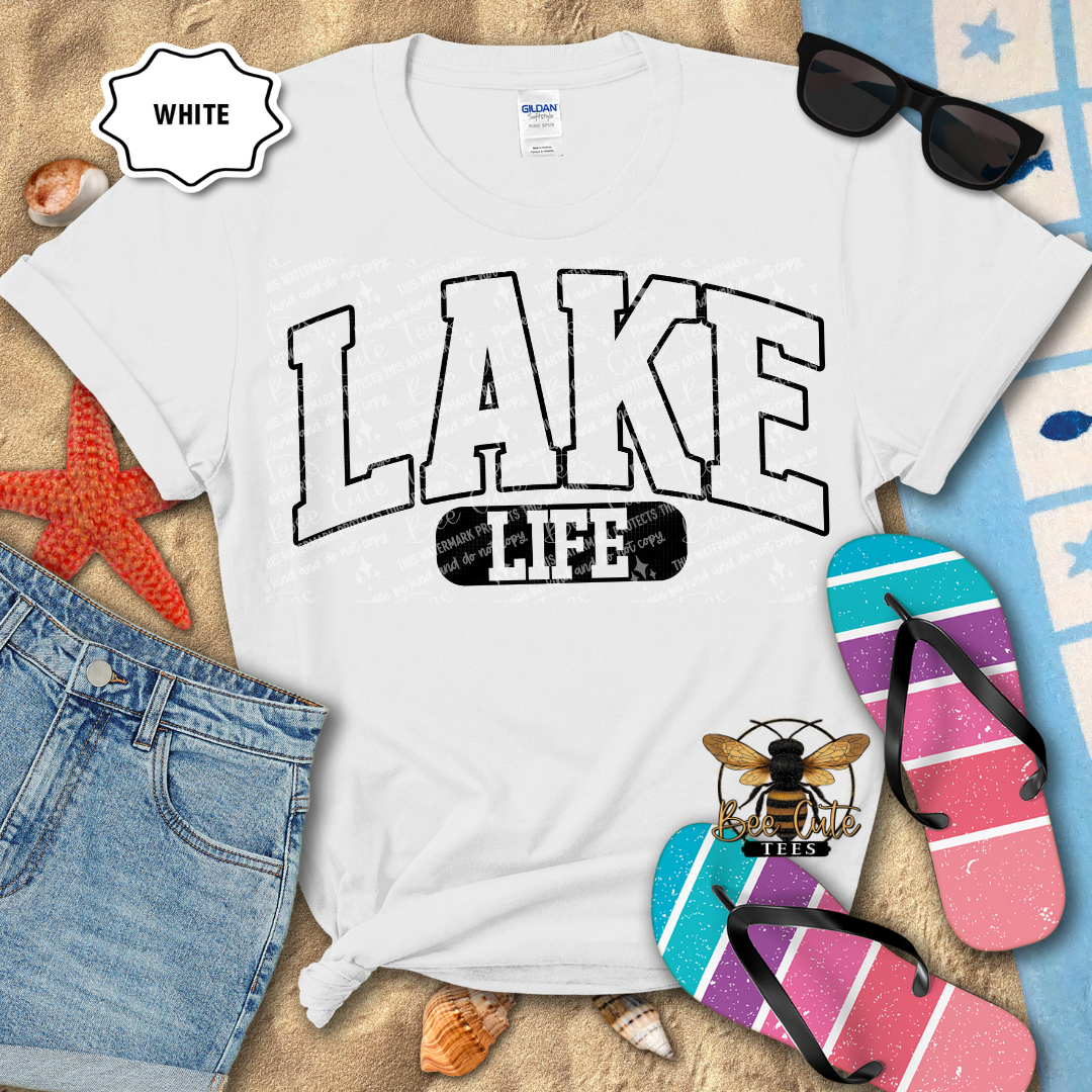 White t-shirt with 'LAKE LIFE' design on a beach setting with sunglasses, flip-flops, and denim shorts.