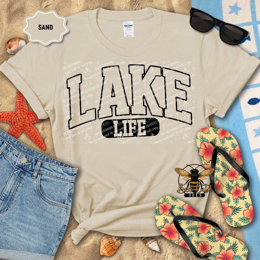 Beige t-shirt with 'Lake Life' print on a sandy background with flip-flops and sunglasses.