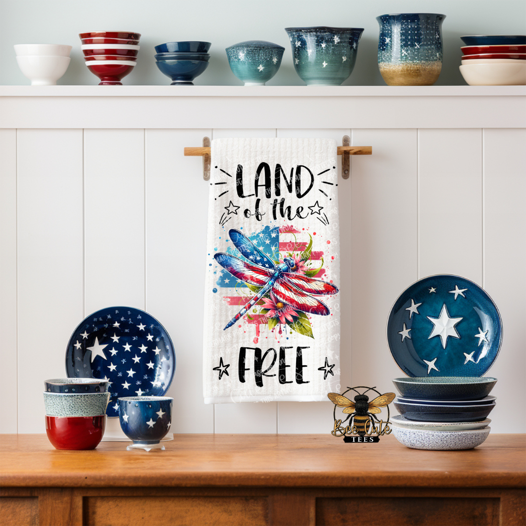 White kitchen towel hanging on a rack in a farmhouse-style kitchen, featuring a red, white, and blue dragonfly over an American flag background with the phrase “Land of the Free,” surrounded by patriotic dishes and decor on a wooden counter.
