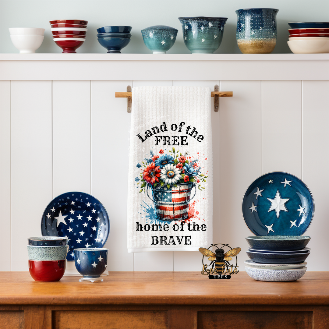 White kitchen towel hanging on a wall hook, featuring a patriotic design with a red, white, and blue flower arrangement in a bucket and the phrase “Land of the Free, home of the Brave,” surrounded by decorative dishes and bowls on a shelf and countertop.
