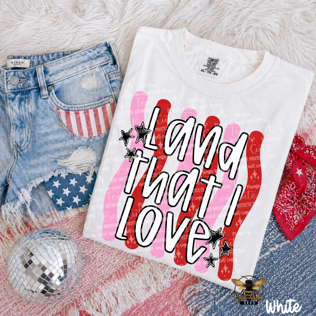 White t-shirt with 'Land That I Love' design on a textured surface with denim shorts and disco ball.