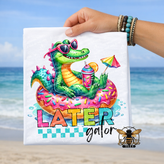 Colorful t-shirt with a cartoon alligator on an inflatable donut, text 'LATER gator' held by a person at the beach.