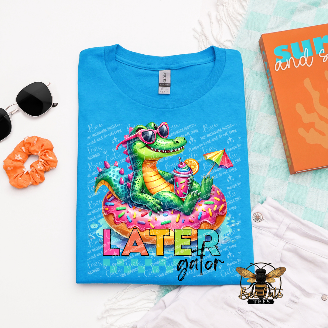 Blue t-shirt with a colorful alligator graphic and 'LATER gator' text, surrounded by sunglasses, scrunchie, and notebook.