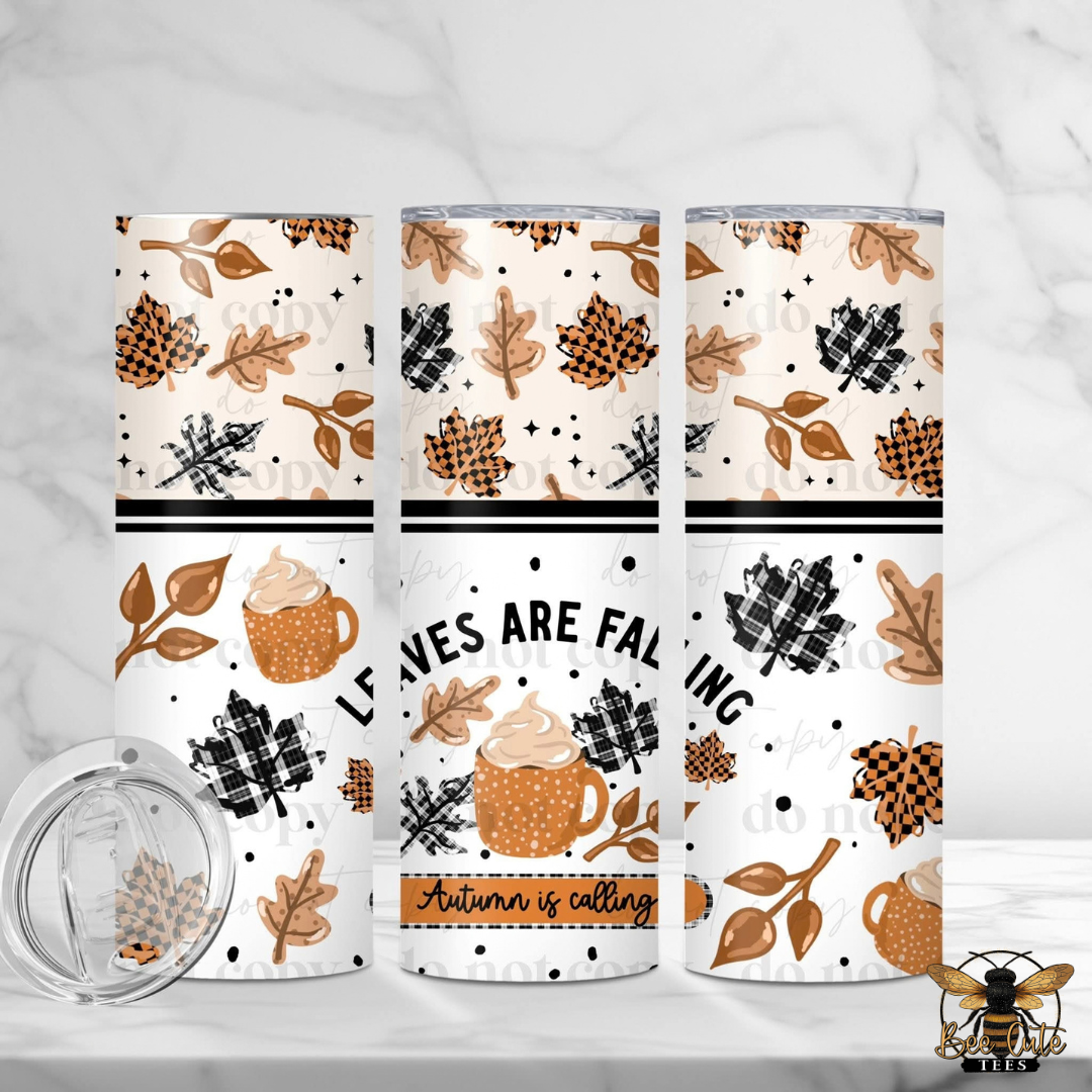 Three insulated tumblers with a fall-themed pattern are displayed on a white marble surface. The pattern features autumn leaves, pinecones, plaid motifs, and a hot cocoa cup, with one tumbler showing the text 'LEAVES ARE FALLING' and 'Autumn is calling'.