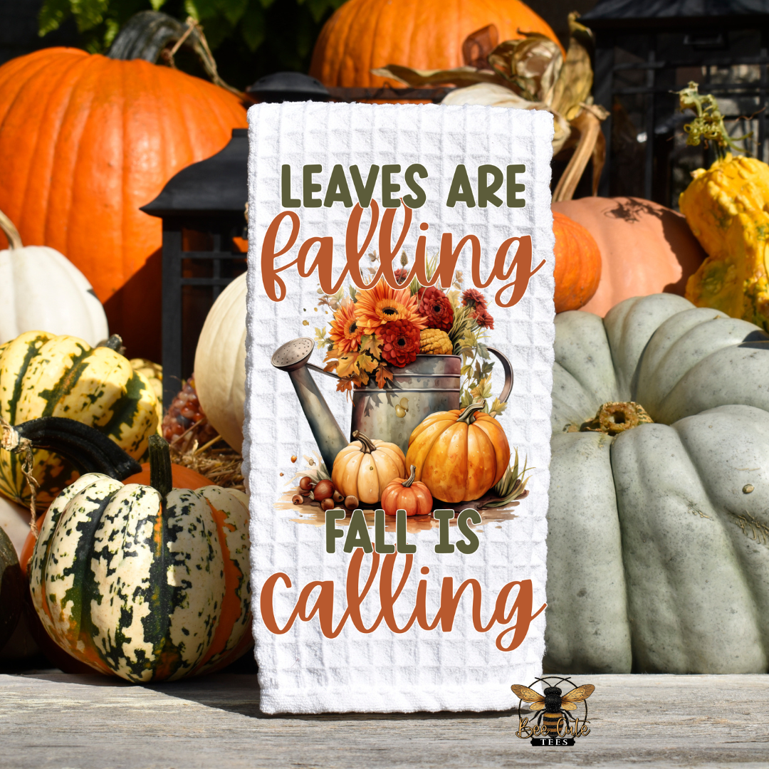 A white waffle-knit rectangular tea towel or sign with autumn-themed text reading 'LEAVES ARE falling FALL IS calling' and a floral watering can illustration, displayed outdoors among pumpkins and gourds.