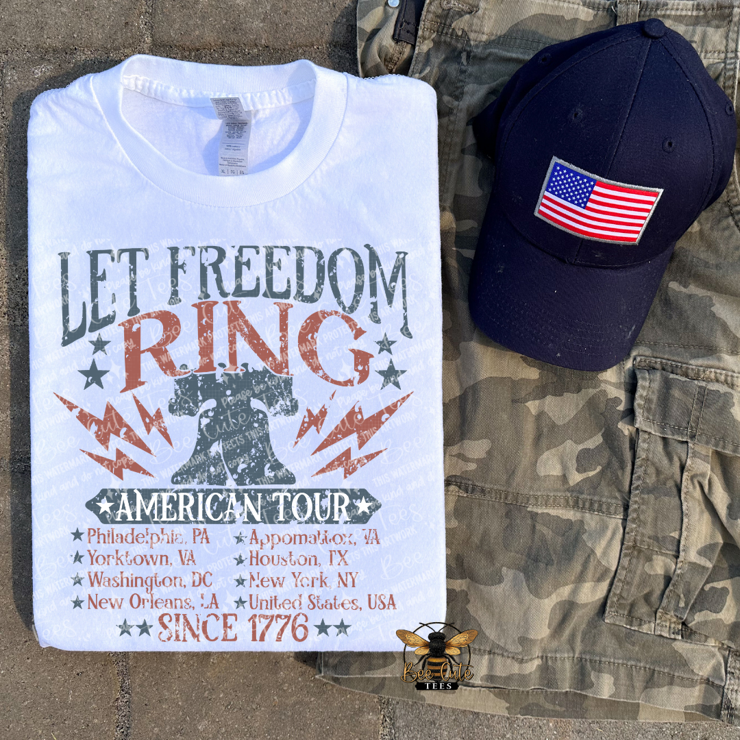 T-shirt with 'Let Freedom Ring' design, cap with American flag, and camouflage pants on a textured surface.