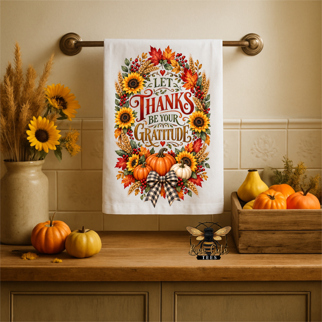 A white kitchen towel with a colorful autumn-themed graphic reading 'LET THANKS BE YOUR GRATITUDE' hangs on a brass towel bar above a wooden counter. The design features sunflowers, pumpkins, fall leaves, and a plaid bow.