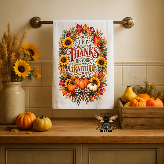 A white kitchen towel with a colorful autumn-themed graphic reading 'LET THANKS BE YOUR GRATITUDE' hangs on a brass towel bar above a wooden counter. The design features sunflowers, pumpkins, fall leaves, and a plaid bow.