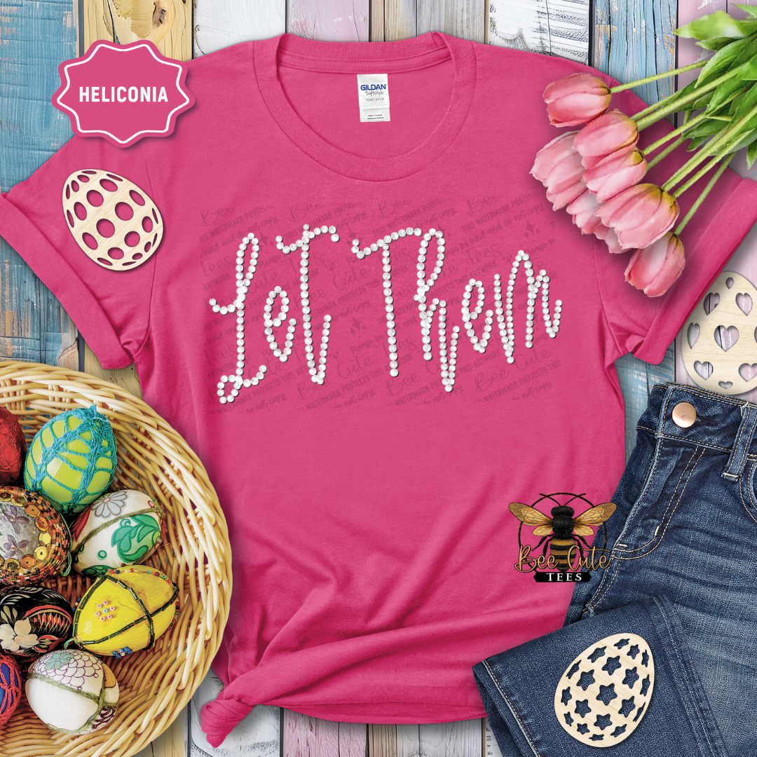 Pink t-shirt with 'Let Them' text, surrounded by Easter decorations on a wooden surface.