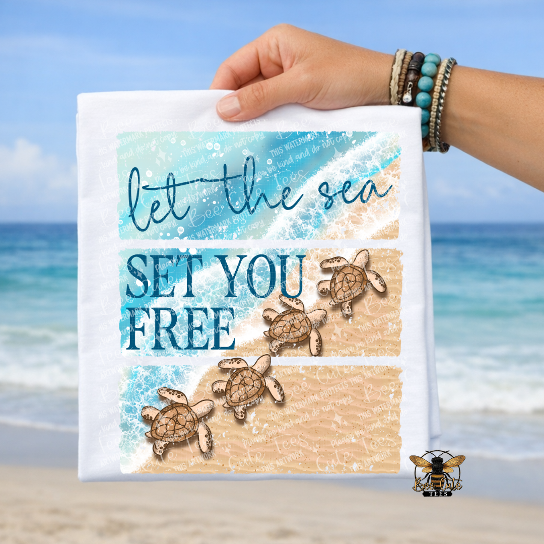 Hand holding a white t-shirt with sea turtles and 'let the sea set you free' text against a beach background