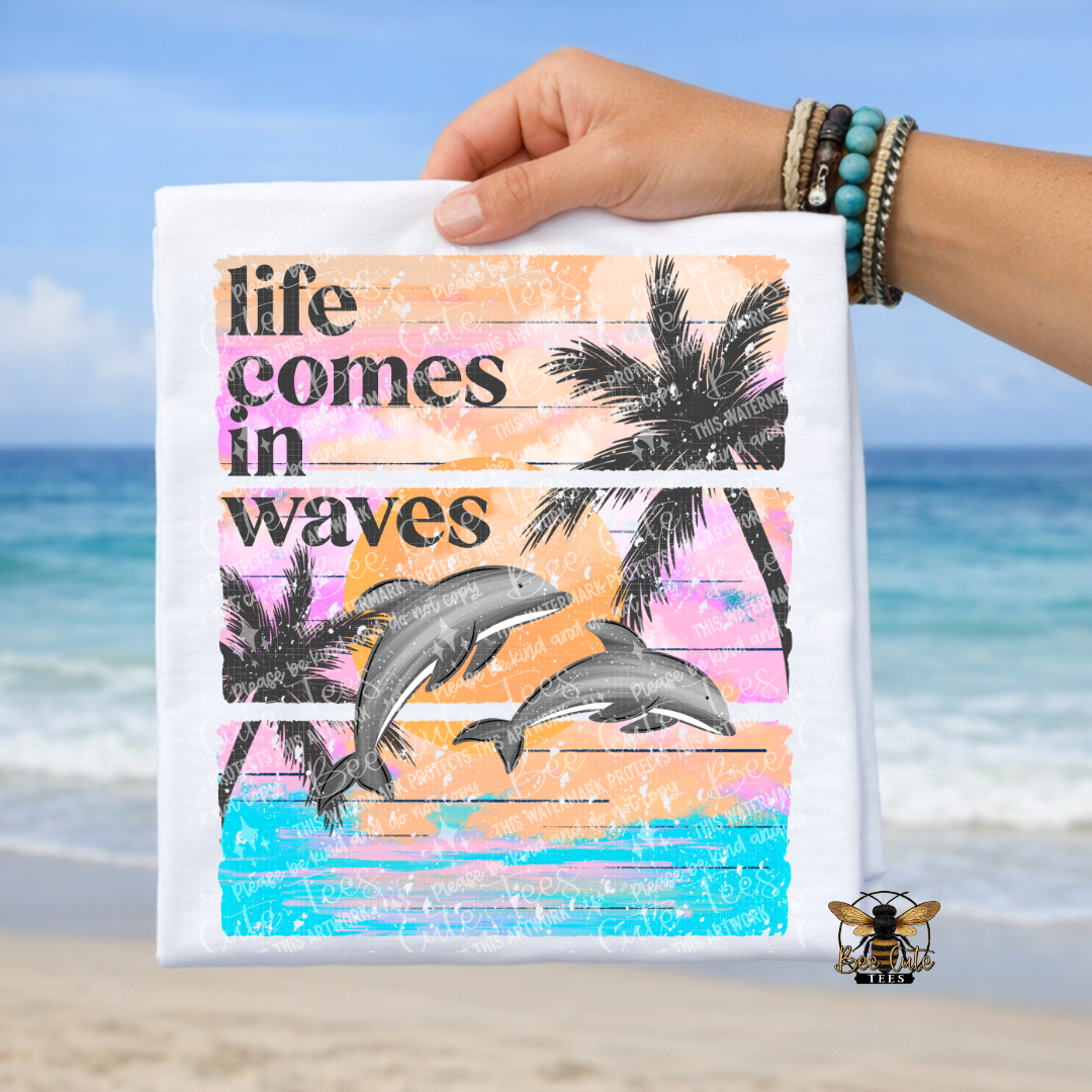 Hand holding a white t-shirt with 'life comes in waves' text and dolphins design against a beach background