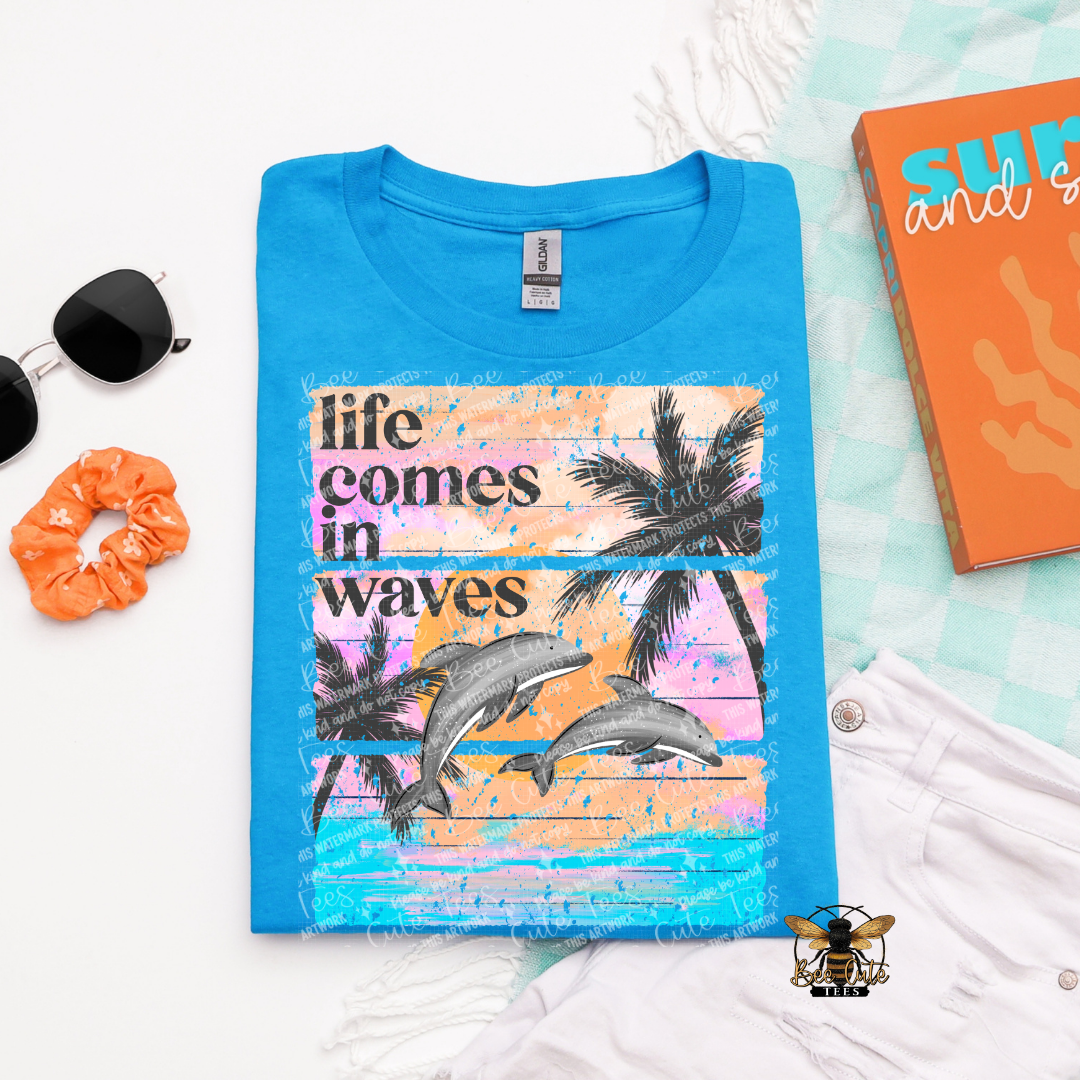 Blue t-shirt with 'life comes in waves' design featuring dolphins and palm trees, placed on a light blue surface with accessories.