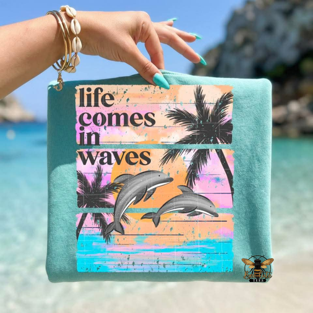 Hand holding a colorful t-shirt with 'life comes in waves' text and dolphins design against a beach background.