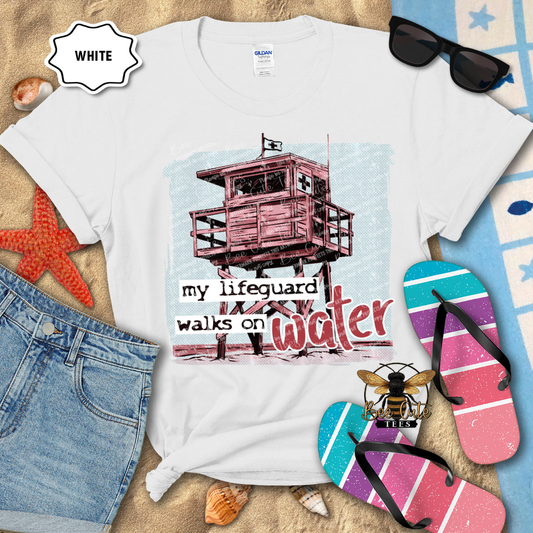 White t-shirt with a lifeguard tower graphic and text, surrounded by beach accessories.