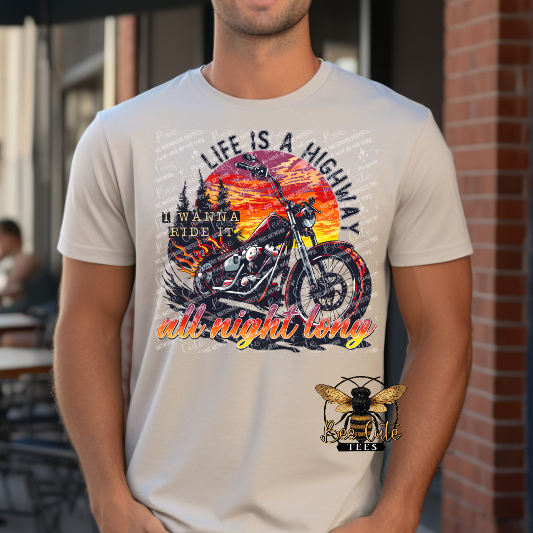 Man wearing a light beige graphic tee featuring a black motorcycle against a vibrant sunset with the phrase “Life Is a Highway” in colorful retro-style lettering.
