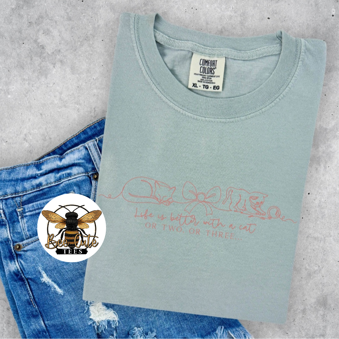 A folded sage green t-shirt with a pink graphic print of a cat and the text 'Life is better with a cat OR TWO OR THREE' laid on a gray concrete background next to blue jeans. A circular bee logo sticker is on the left side of the image.