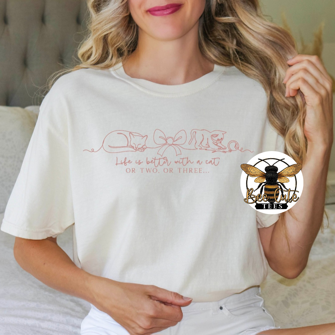 A woman wearing a white-colored t-shirt with a pink graphic that reads 'Life is better with a cat OR TWO OR THREE.