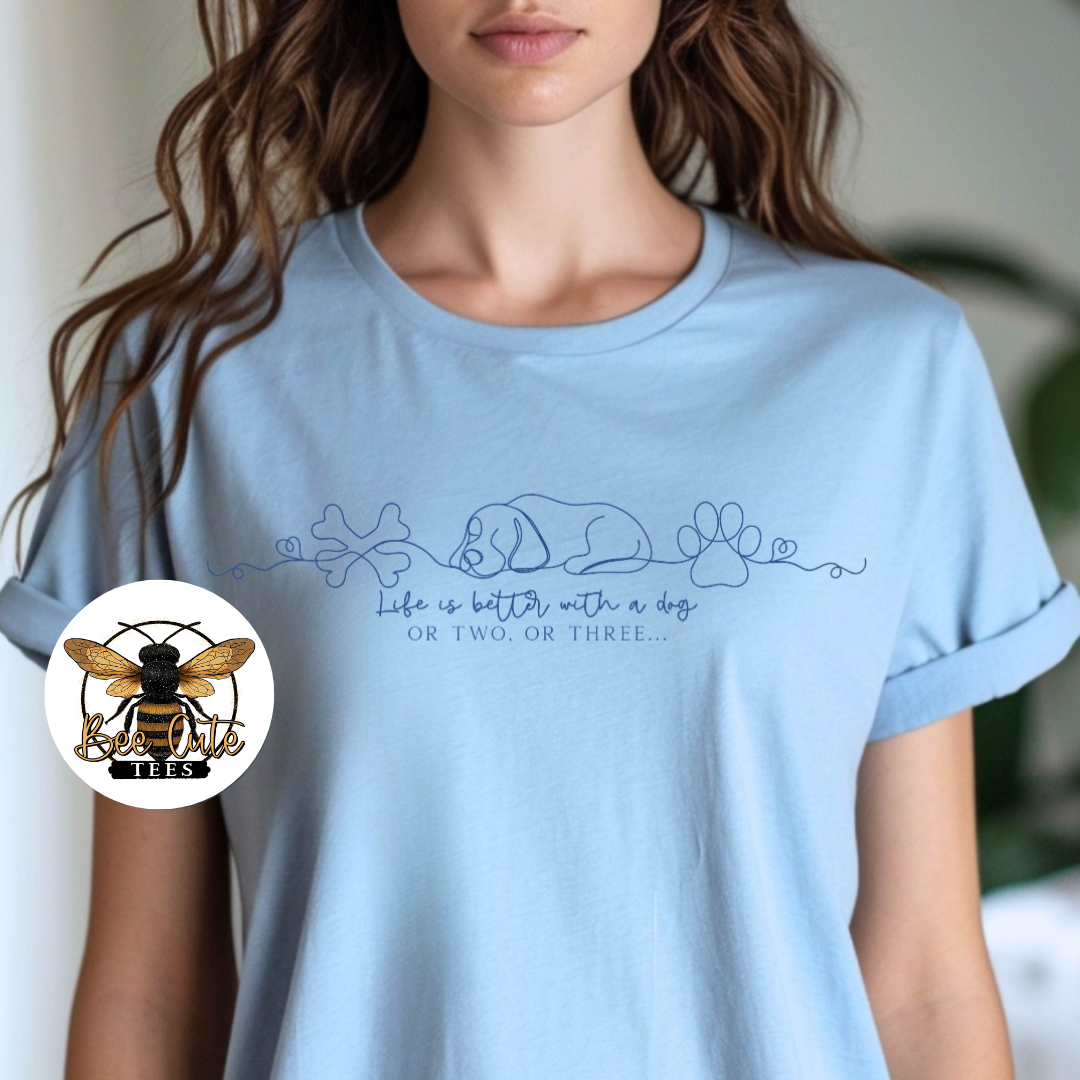 A woman wearing a light blue crewneck t-shirt with a dark blue line art graphic of a sleeping dog, a paw print, and the text 'Life is better with a dog OR TWO, OR THREE.