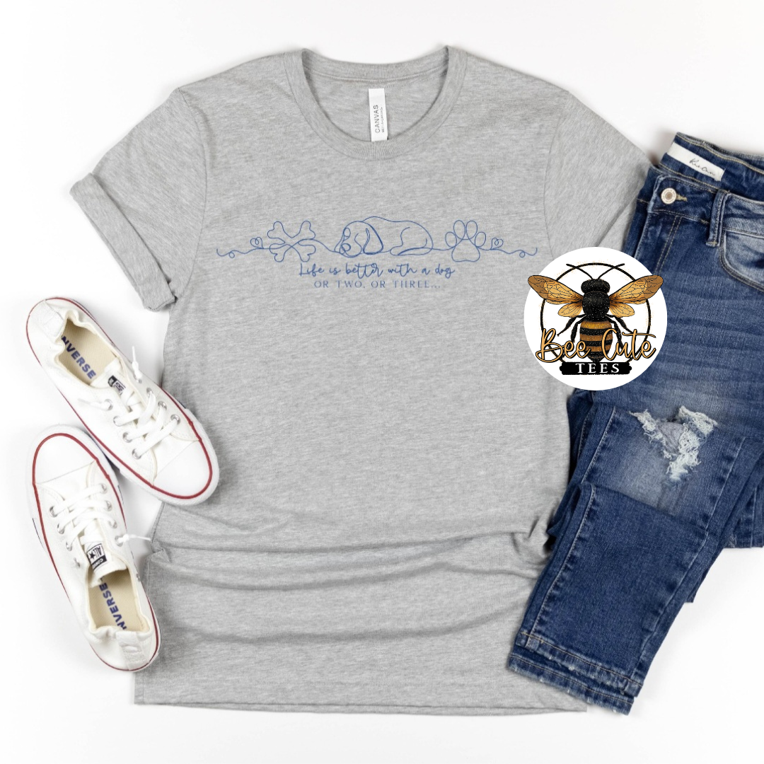 A heather gray graphic t-shirt featuring blue line art of a dog, cat, and paw prints with the text 'life is best with a dog OR two or three', styled in a flat lay with white sneakers, blue jeans, and a circular bee logo sticker.