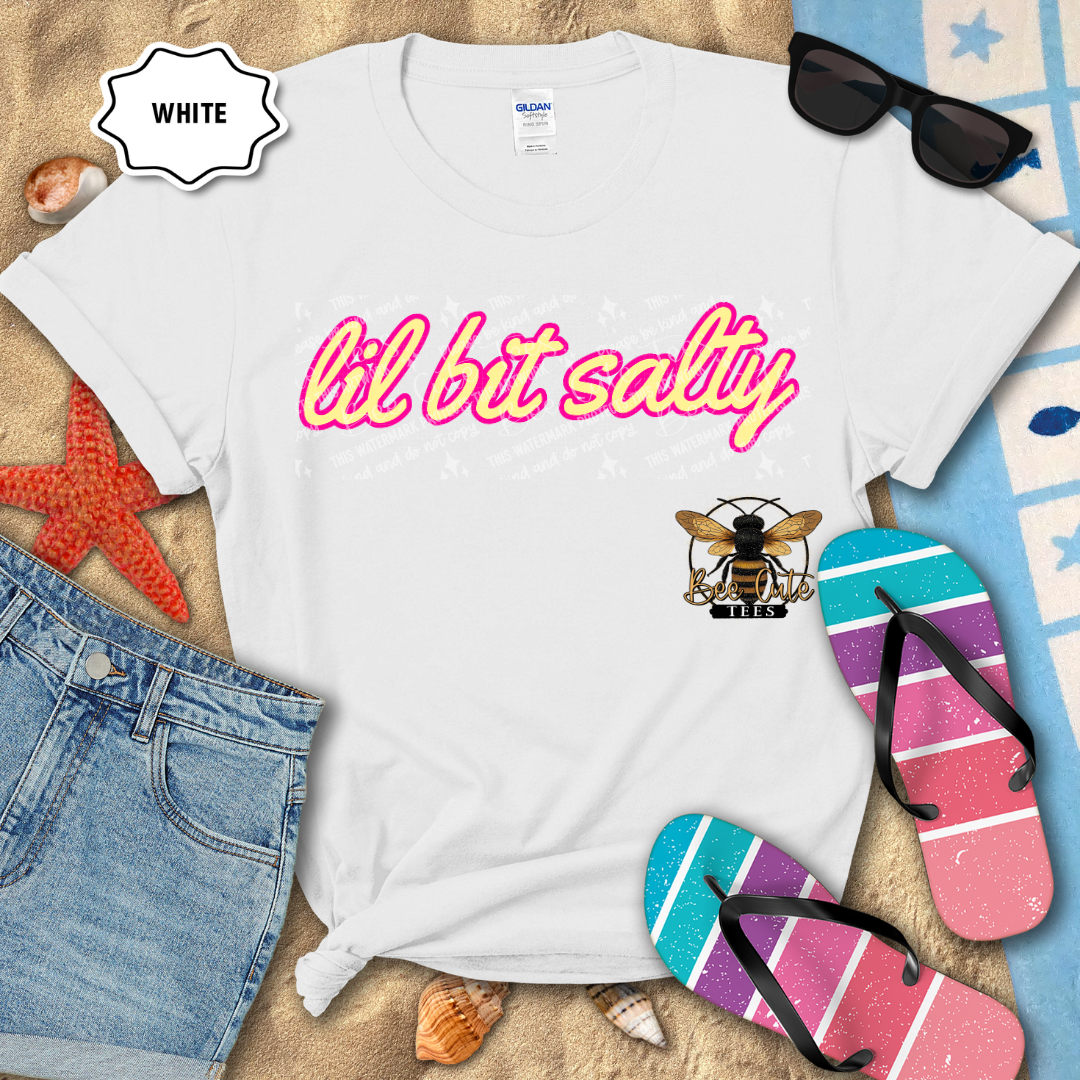 White t-shirt with 'lil bit salty' text and bee logo on a beach-themed background with sunglasses, flip-flops, and denim shorts.