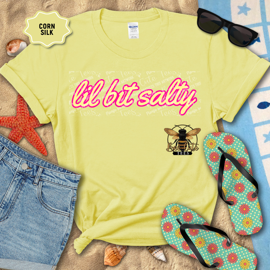 Yellow t-shirt with 'lil bit salty' text and bee logo on a beach setting with sunglasses, flip-flops, and starfish.