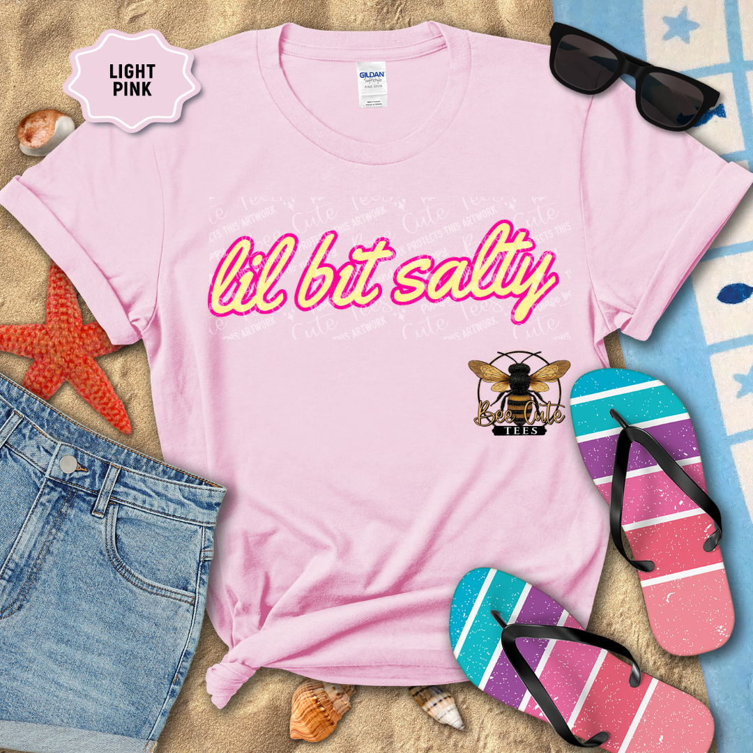 Light pink t-shirt with 'lil bit salty' text and bee logo, surrounded by beach accessories.