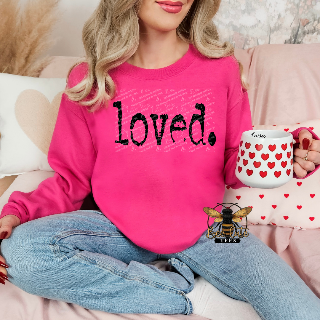 Person wearing a pink sweatshirt with 'loved' text, holding a mug with heart patterns, sitting on a couch.