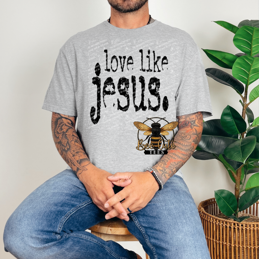 Person wearing a gray t-shirt with 'love like jesus' text and a bee logo, sitting next to a plant.