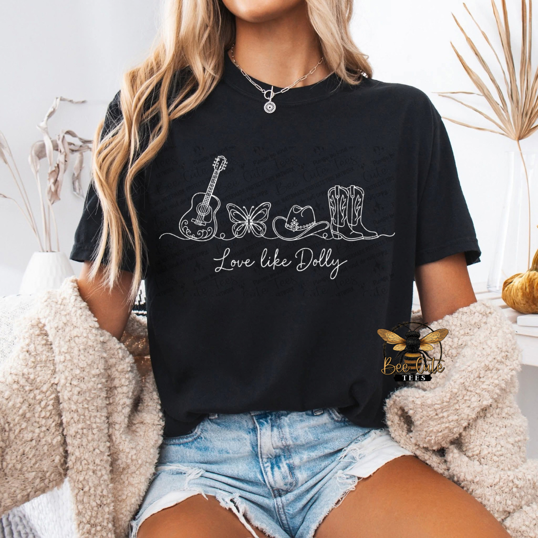 A woman wearing a black graphic t-shirt with a white line art design of a guitar, butterfly, cowboy hat, boots, and the text 'Love like Dolly'. She is also wearing denim shorts and a beige sherpa jacket, with a 'Love like Dolly' brand logo overlay on the bottom right.