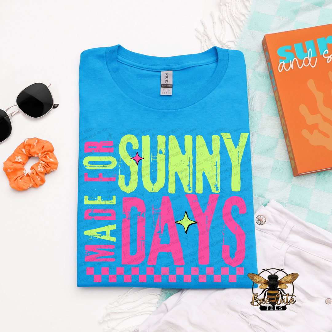 Blue t-shirt with 'Made for Sunny Days' text on a white surface with accessories.