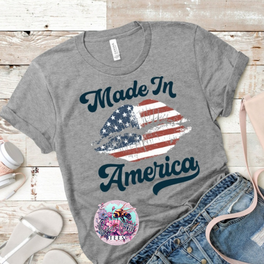 Gray t-shirt featuring a “Made in America” graphic with an American flag lip design, styled flat lay with denim shorts and accessories.
