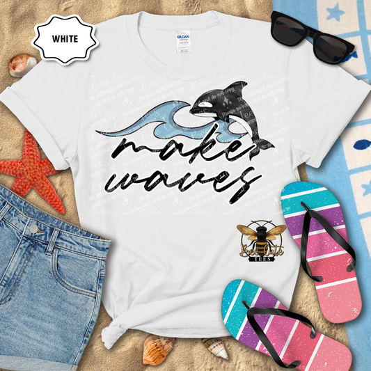 White t-shirt with whale design and 'make waves' text, surrounded by beach items on sand.