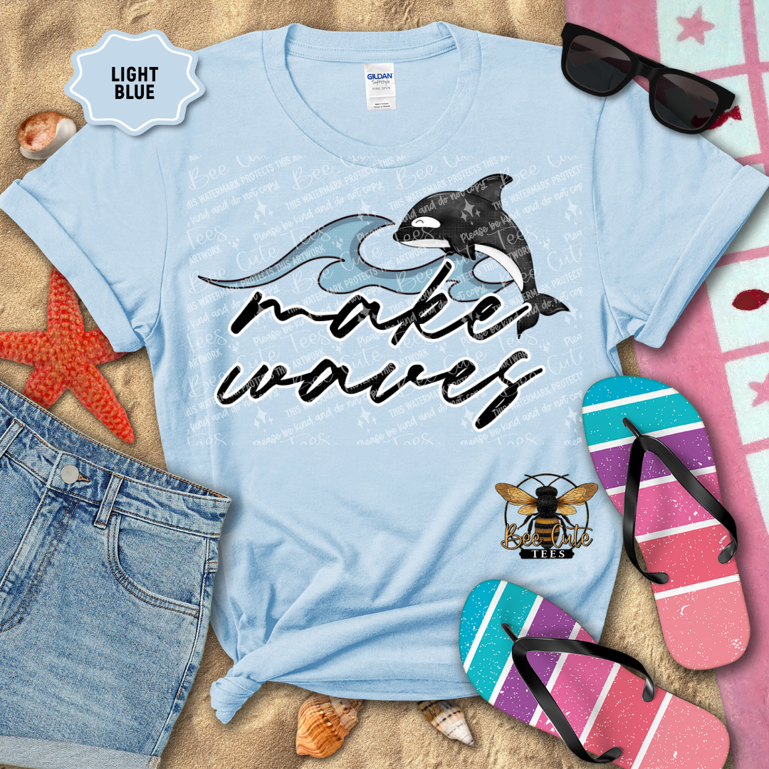 Light blue t-shirt with dolphins and 'make waves' text, surrounded by beach accessories on sand.