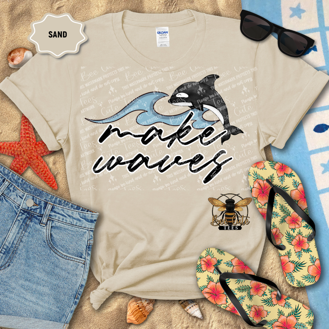 Beige t-shirt with 'make waves' and dolphin design, surrounded by beach accessories on sand.