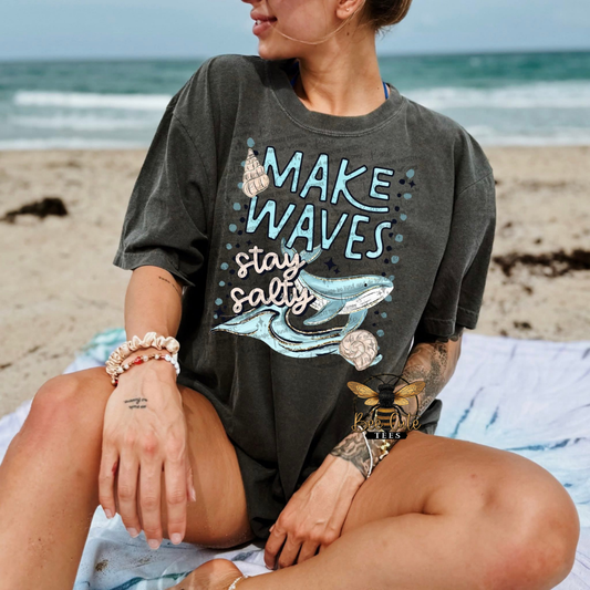 Person wearing a t-shirt with 'Make Waves, Stay Salty' design on a beach.