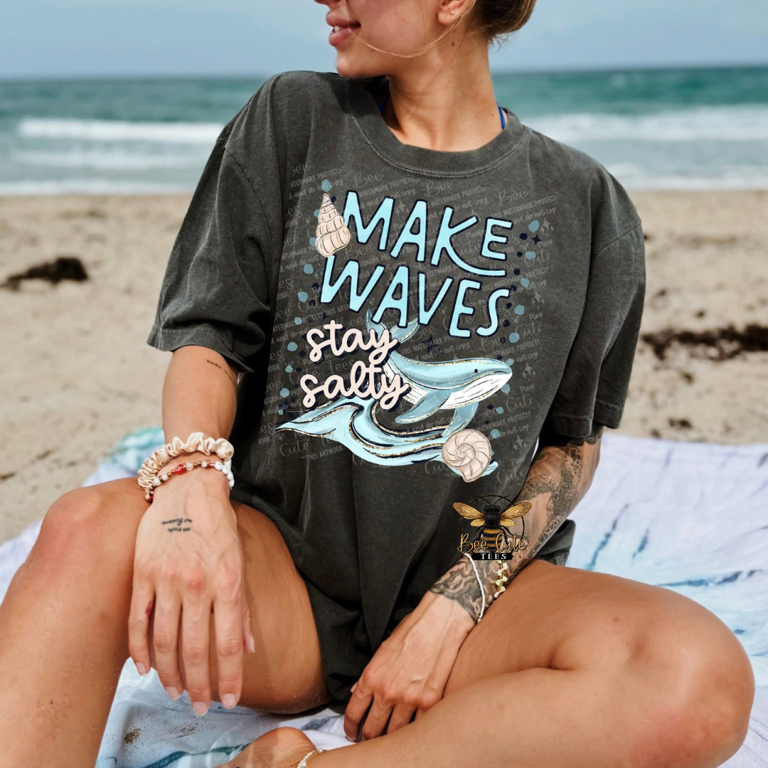 Person wearing a t-shirt with 'Make Waves, Stay Salty' design on a beach.