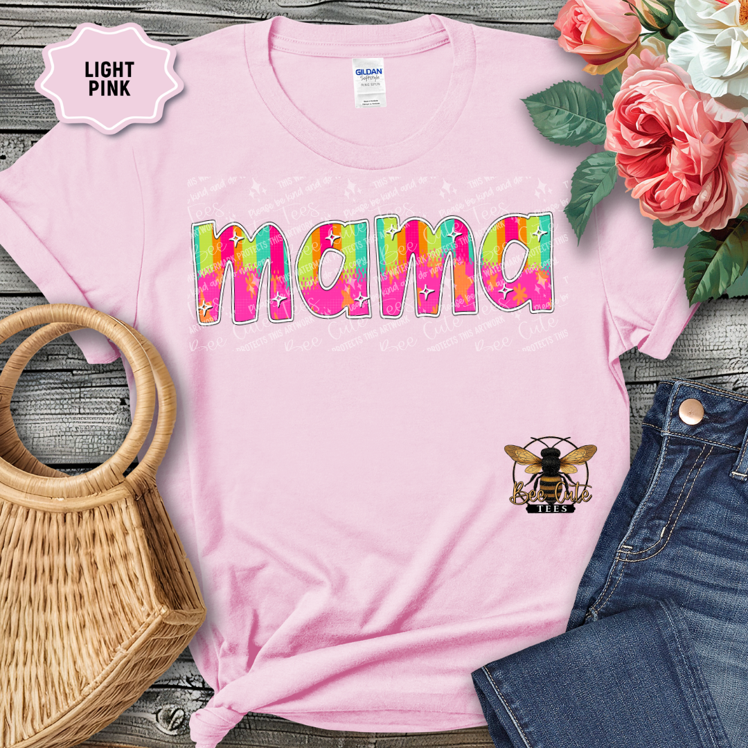 Light pink t-shirt with colorful 'mama' text and bee graphic, placed on a wooden surface with flowers and denim jeans.