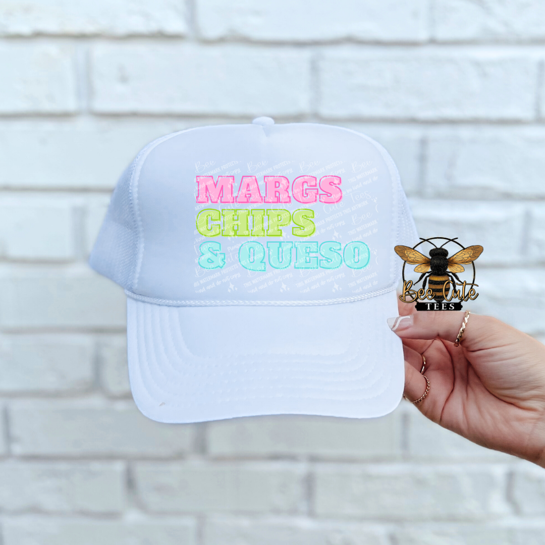 White cap with colorful text 'MARGS CHIPS & QUESO' held against a white brick wall.