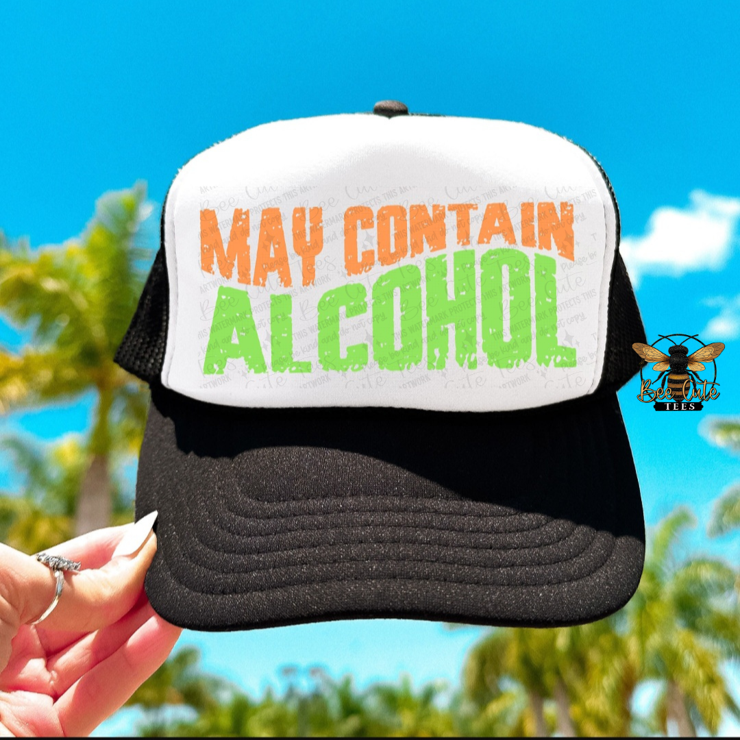 Trucker hat with 'May Contain Alcohol' text held by a hand against a blue sky and greenery background.