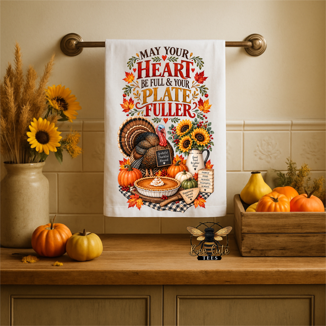 A white kitchen towel with a colorful Thanksgiving-themed graphic hangs from a brass rod on a beige tiled wall above a wooden counter. The graphic features a turkey, pumpkins, sunflowers, and the text 'MAY YOUR HEART BE FULL & YOUR PLATE FULLER.