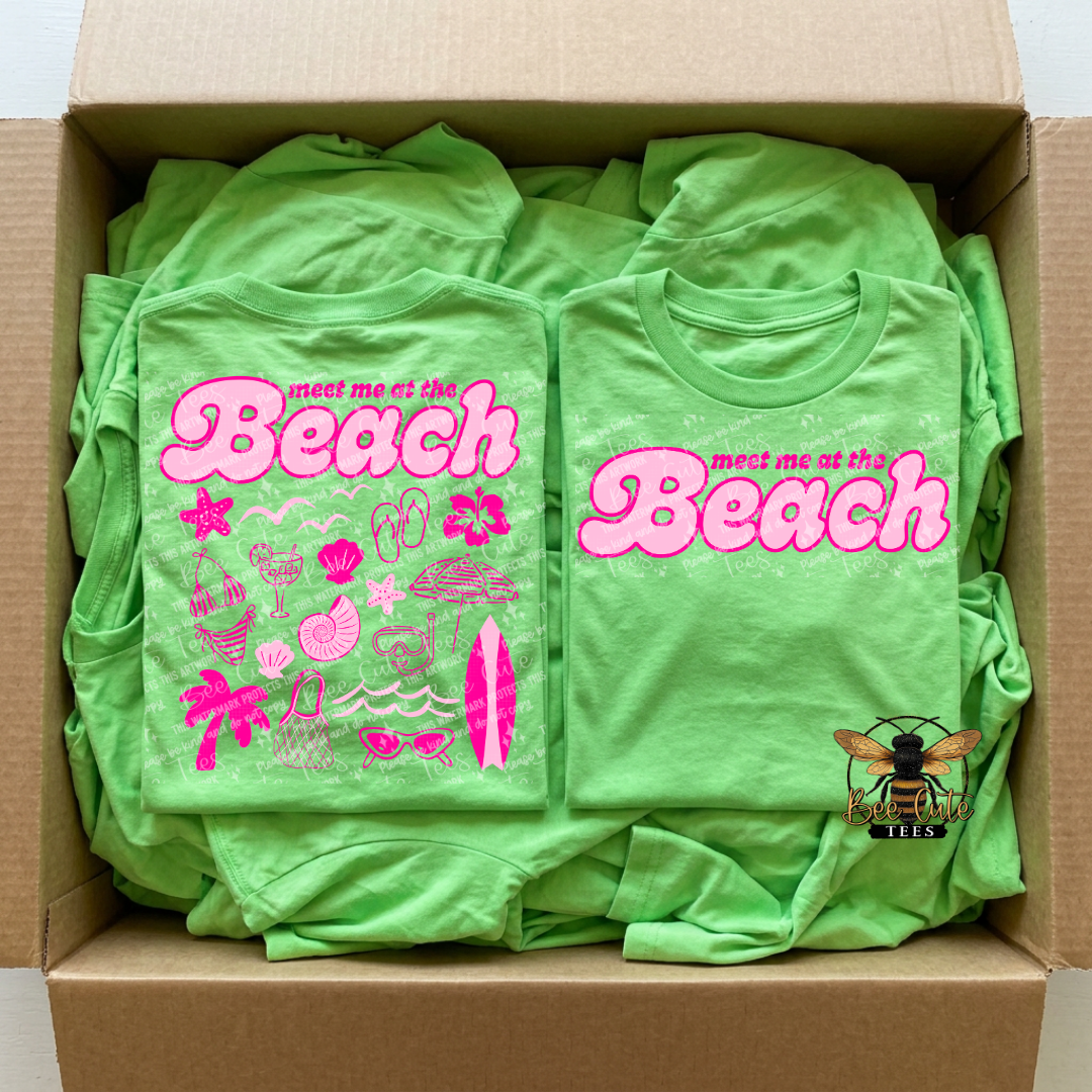 Box of green t-shirts with 'meet me at the Beach' design and pink text.