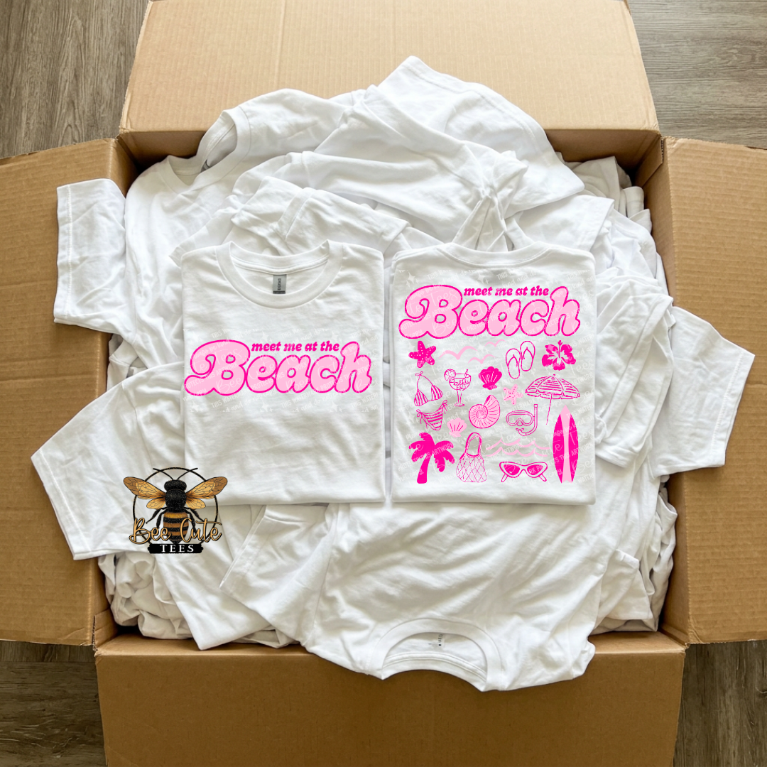 Box of white t-shirts with 'meet me at the Beach' design on a wooden surface