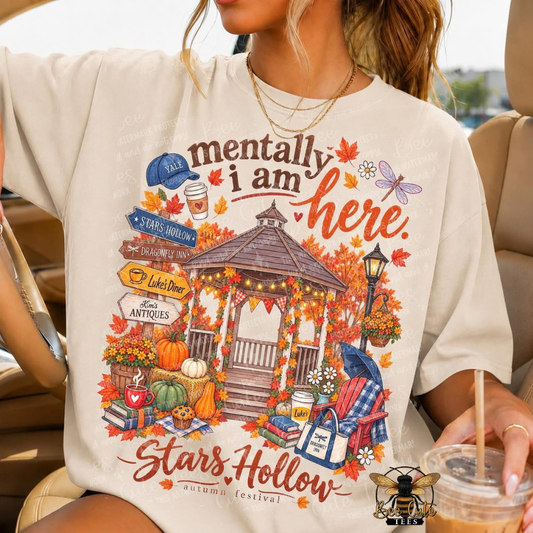 A woman wearing an oversized cream-colored graphic t-shirt in a car. The shirt features a colorful autumn-themed illustration of a gazebo, pumpkins, fall leaves, and the text 'mentally i am here.